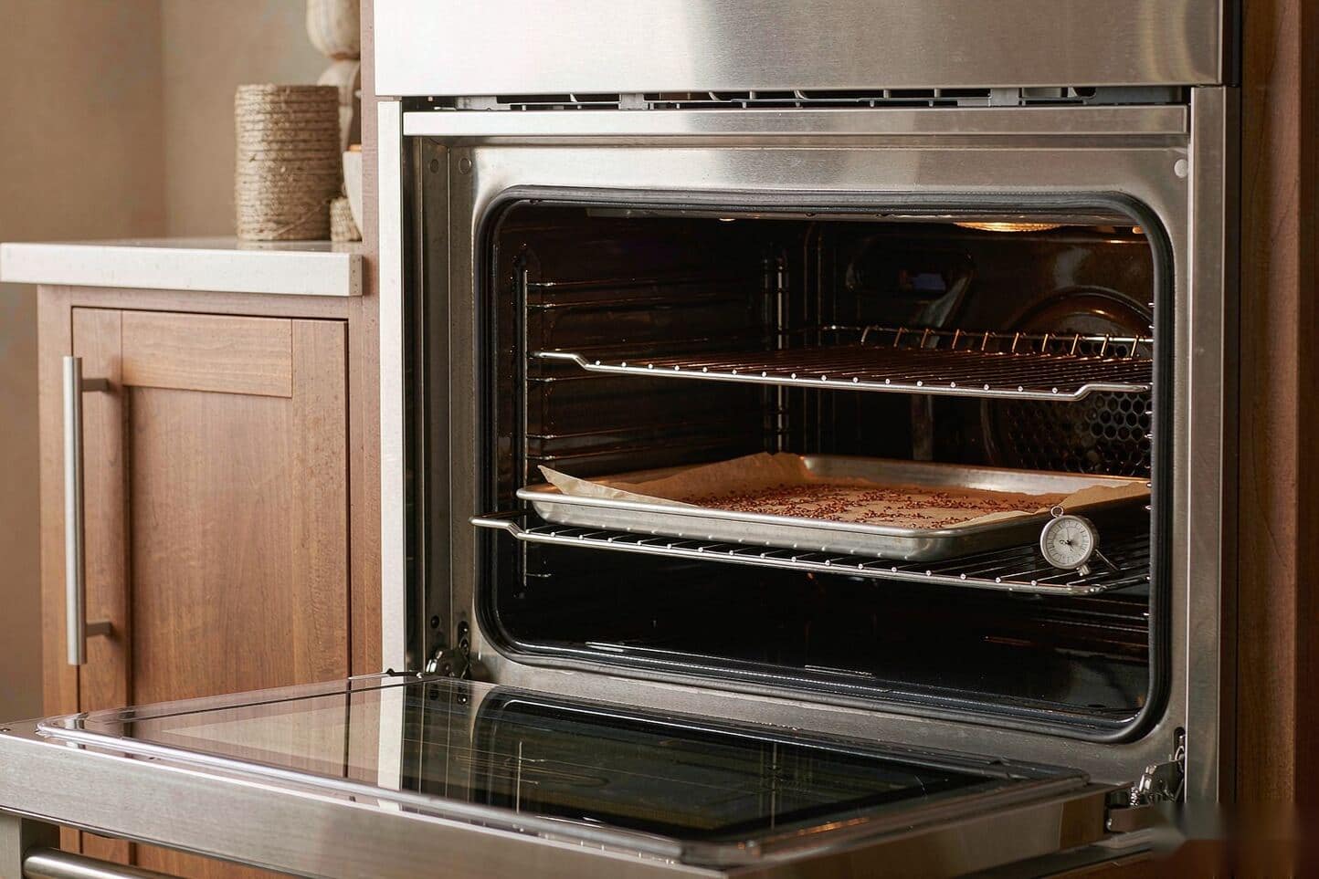 An open oven with a parchment-covered tray of herb and an oven thermometer on the rack.